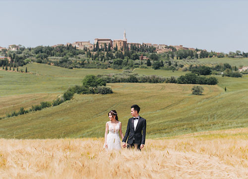bride and groom in wedding clothing pose in field with montepulciano in the background